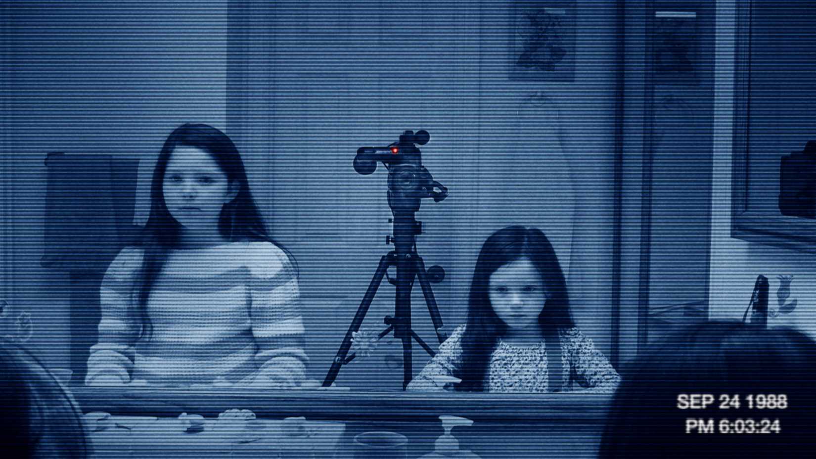 Two young girls with long black hair and jet-black eyes stand in front of a bathroom mirror, a tripod-mounted camera between them, in Paranormal Activity 3
