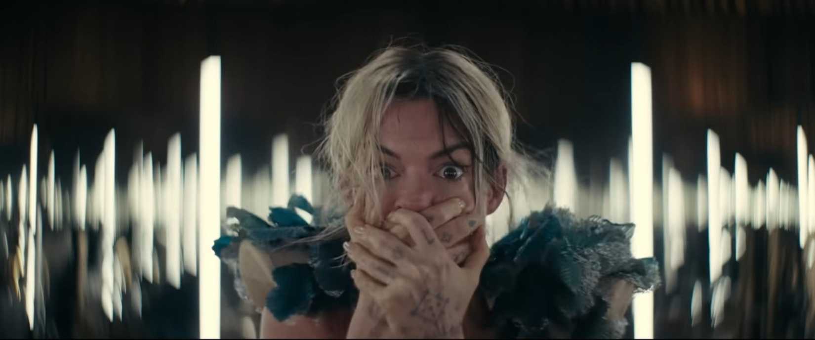 Anne Hathaway, as pop star Mother Mary in the movie of the same name, clasps her hands over her mouth in horror, with stylized lights blurred in the background