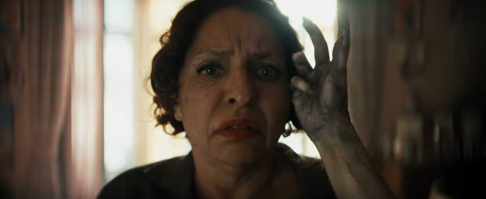 A center-frame close-up of an older woman's face looking uncomfortable as a child's grayish, corpse-like hand caresses the side of her head from off-camera.