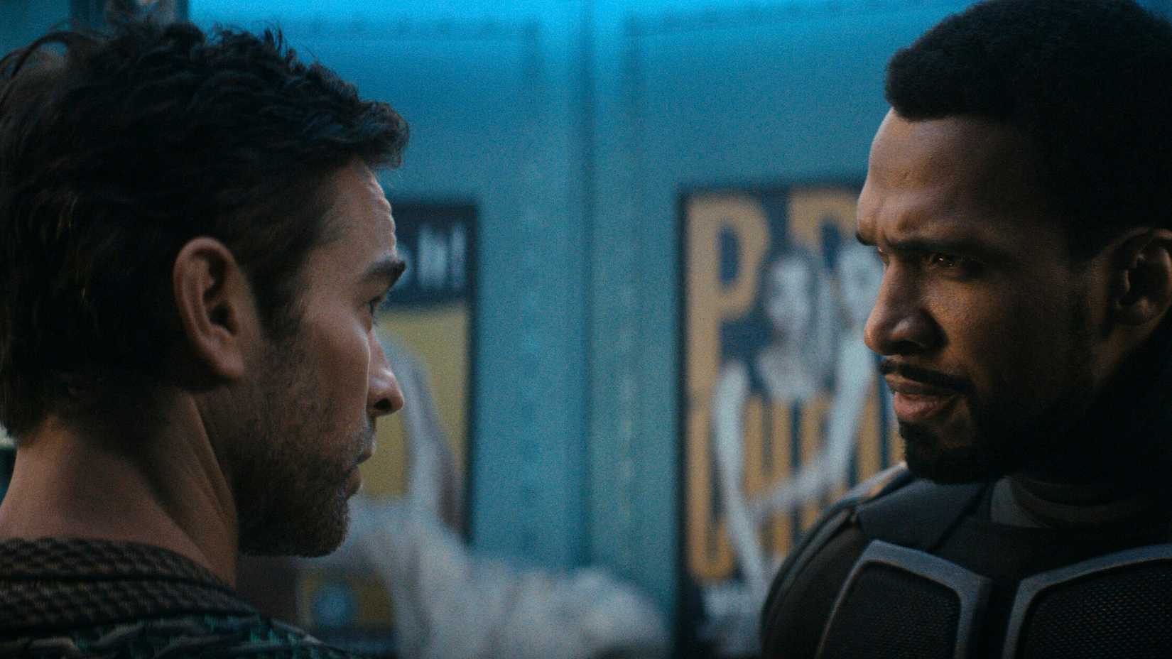 Black Noir (Nathan Mitchell) looks upset with The Deep (Chace Crawford) as they stand in a room filled with posters.