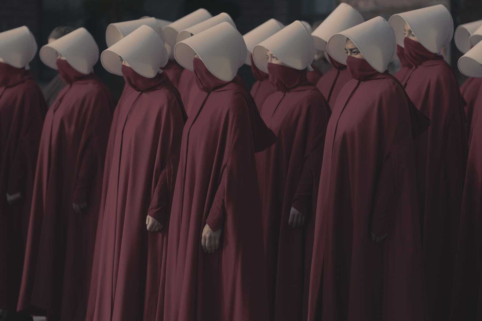 A row of handmaids in The Handmaid's Tale, women in dark red robes and face masks and white wimples, looking sober