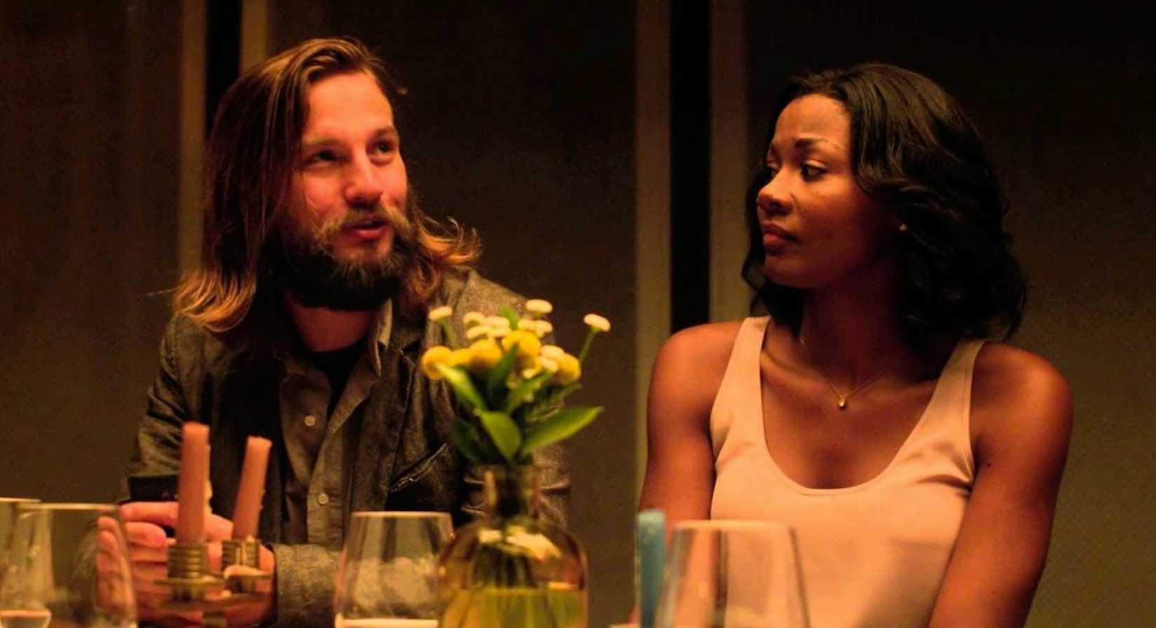 A couple (Logan Marshall-Green and Emayatzy Corinealdi) sit side-by-side at a fancy-looking dinner table, with her looking slightly warily at him, in a scene from The Invitation (2016).