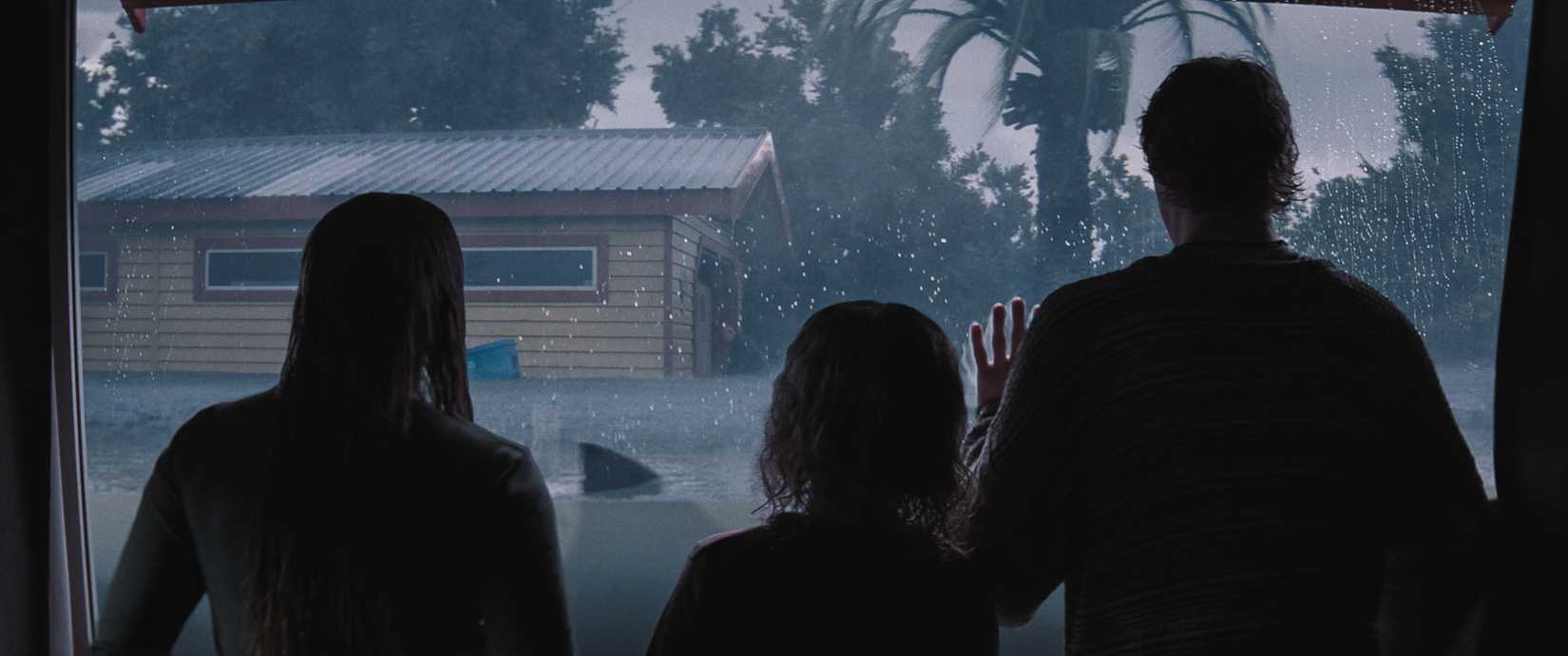 Three children, with their backs to the camera and mostly in shadow, look out of a big glass window to the flooded streets outside, where a shark fin protrudes from the floodwaters, in a scene from the Netflix movie Thrash.
