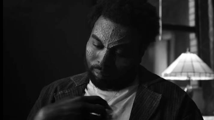 Tombstone (Abraham Popoola) a man with scarification on his forehead holds onto a necklace while looking contemplative in Spider-Noir