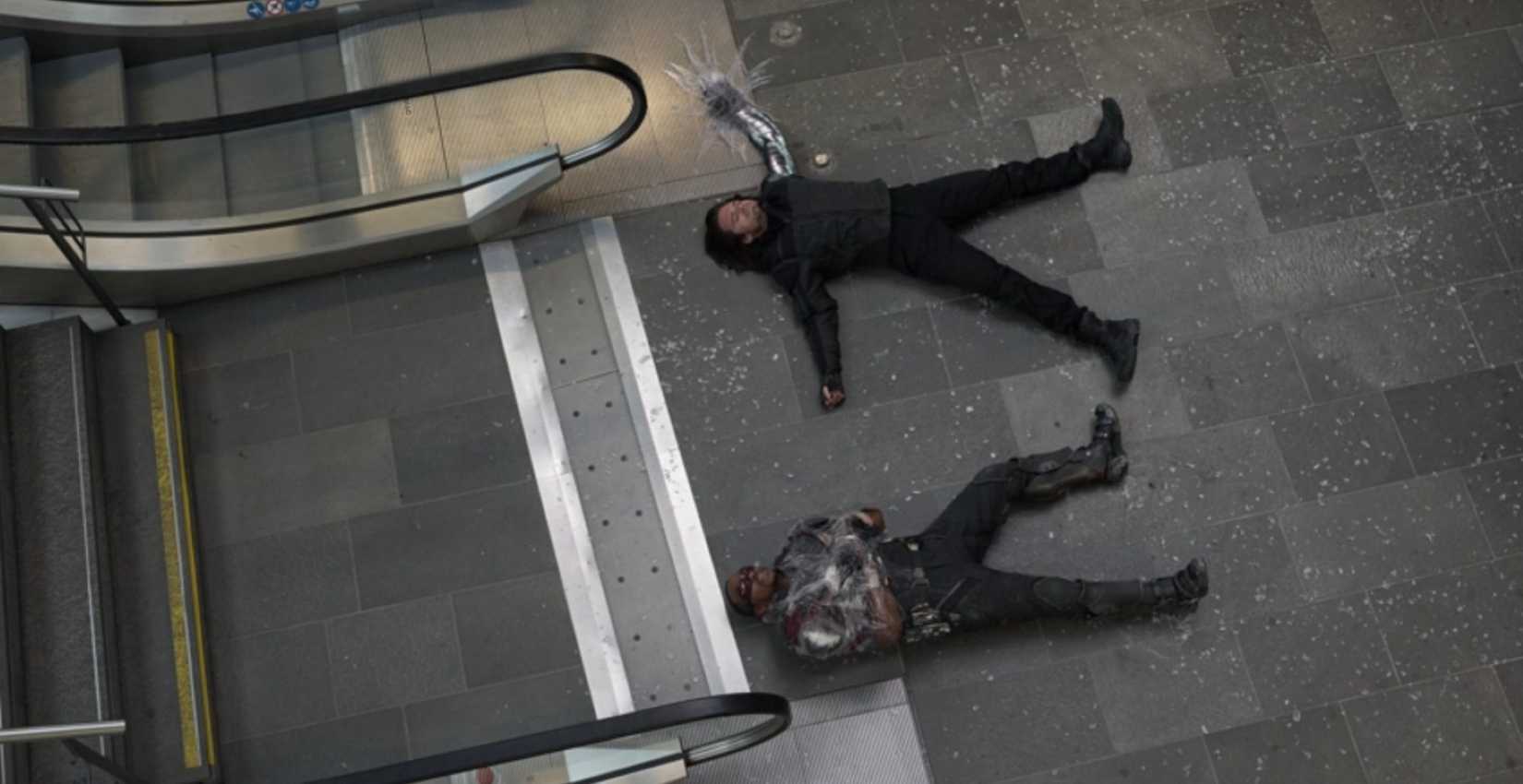 In a desaturated overhead shot from Captain America: Civil War, Bucky (Sebastian Stan) and the Falon (Anthony Mackie) lie on the gray concrete ground in defeat, wearing grayish and navy blue costumes