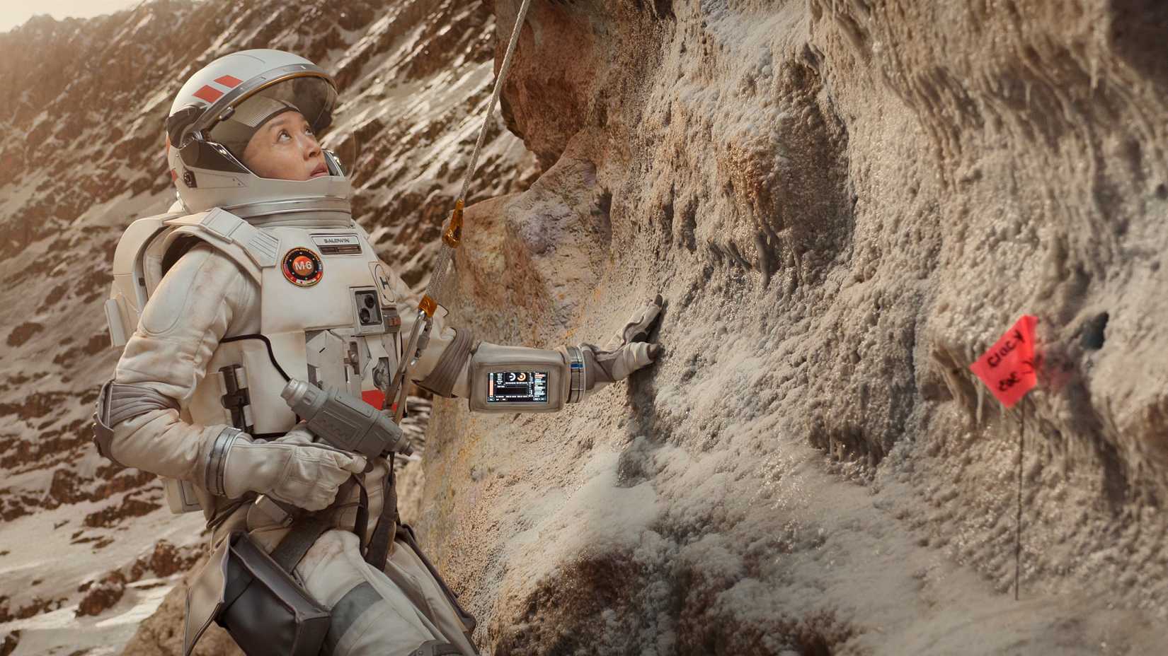 Cynthia Wu wears a spacesuit and holds a tool while looking at a cliff face in For All Mankind season 5.