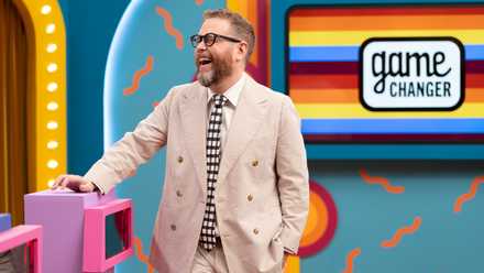 Dropout CEO and Game Changer host Sam Reich, wearing an off-white suit, laughs as he stands by a podium on the set of Dropout's comedy show Game Changer