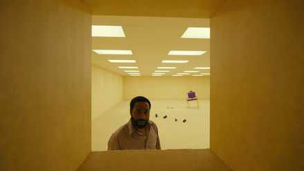 A Black man (Chiwetel Ejiofor) in a button-down shirt stands in a room with yellow wallpaper and a scattering of random objects on the floor behind him and peers through a chest-high, square opening in the wall in Backrooms