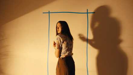 Mary (Renate Reinsve), a thin, pale woman with long black hair, stands with her back to the camera, looking nervously back over her shoulder, in front of a tape outline of a doorway on a blank wall in Backrooms