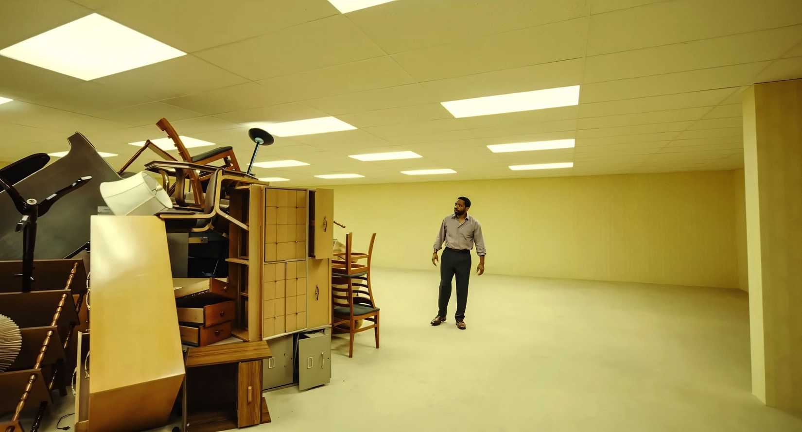 A Black man (Chiwetel Ejiofor) in a button-down shirt and black slacks stands in a huge room with the walls, floor, and ceiling all painted the same sallow yellow. He's examining a huge pile of jumbled furniture, the only thing in the room.