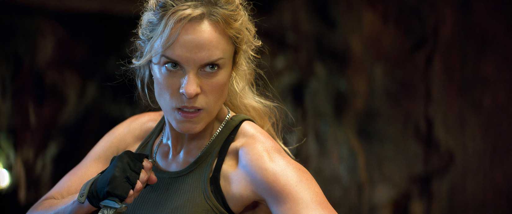 Sonya Blade (Jessica McNamee) in Mortal Kombat 2, a blonde woman in a sleeveless green T-shirt, fists clenched, looking determined.