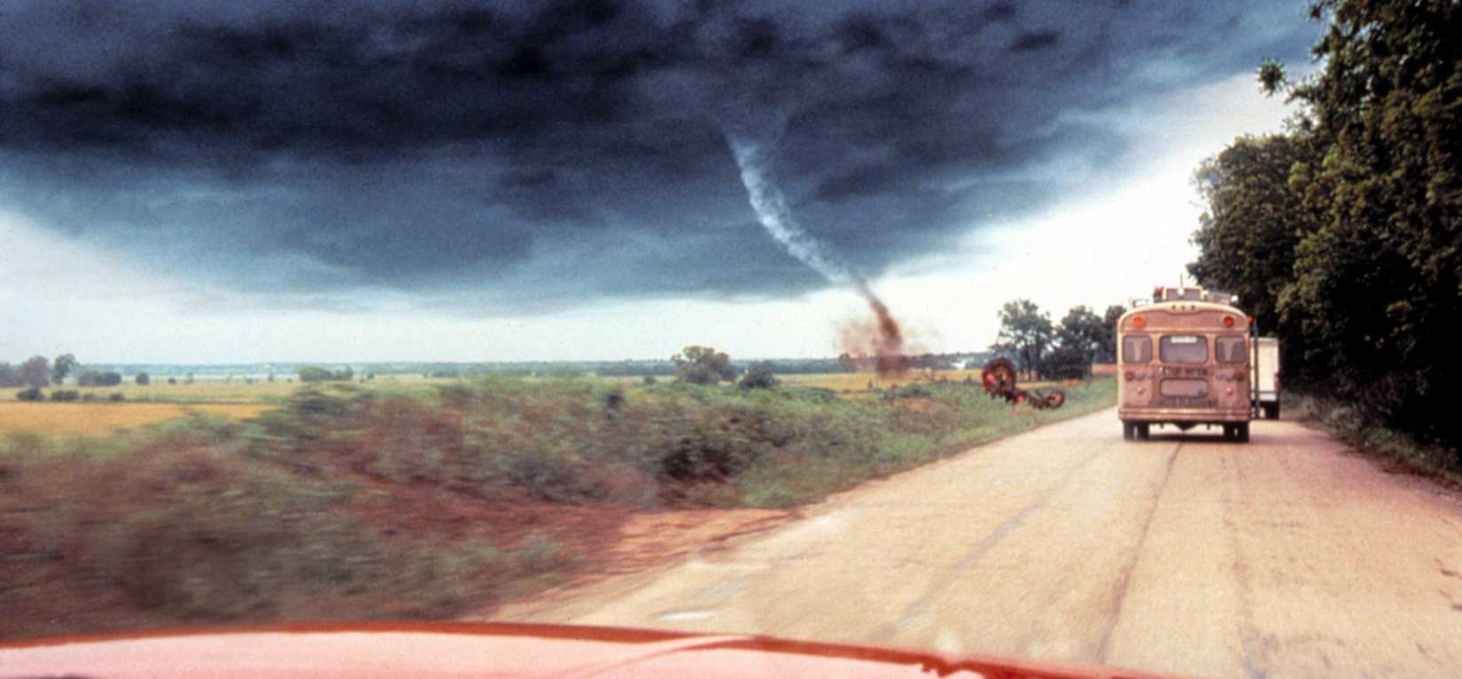 A widescreen shot showing a tornado in the background with an old bus seen over the hood of a truck