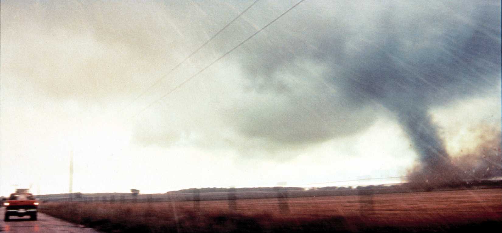 A widescreen shot of a tornado on the right and a truck on the left in a wide open plain