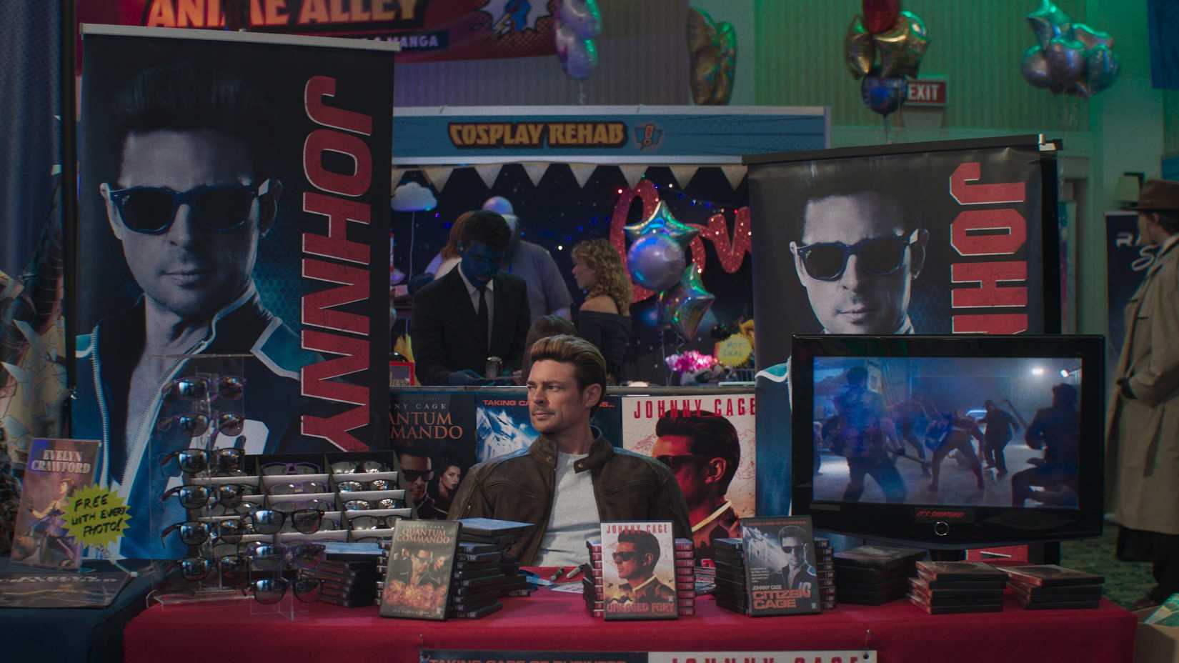 Johnny Cage (Karl Urban) sits at a booth at a convention surrounded by DVDs and sunglasses in Mortal Kombat 2.
