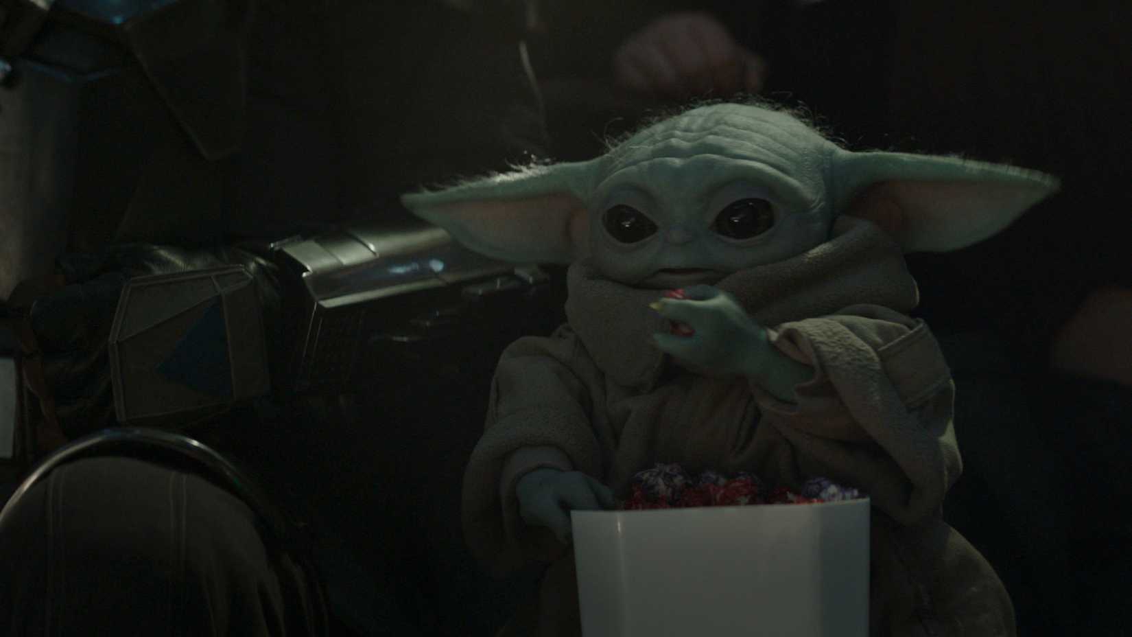 Grogu snacks on popcorn while sitting next to Din Djarin at a gladiator match in The Mandalorian and Grogu.
