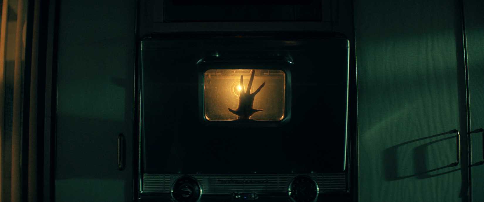 A dark, five-fingered hand stretches out from behind an oven. It is backlit by an orange light.