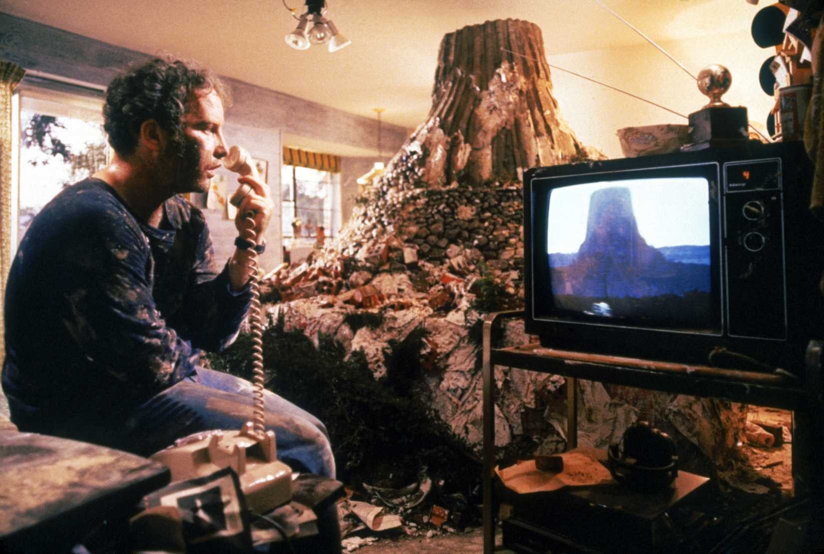 Richard Dreyfuss looks at an image of a mountain on his TV, with a huge mud model of it behind him
