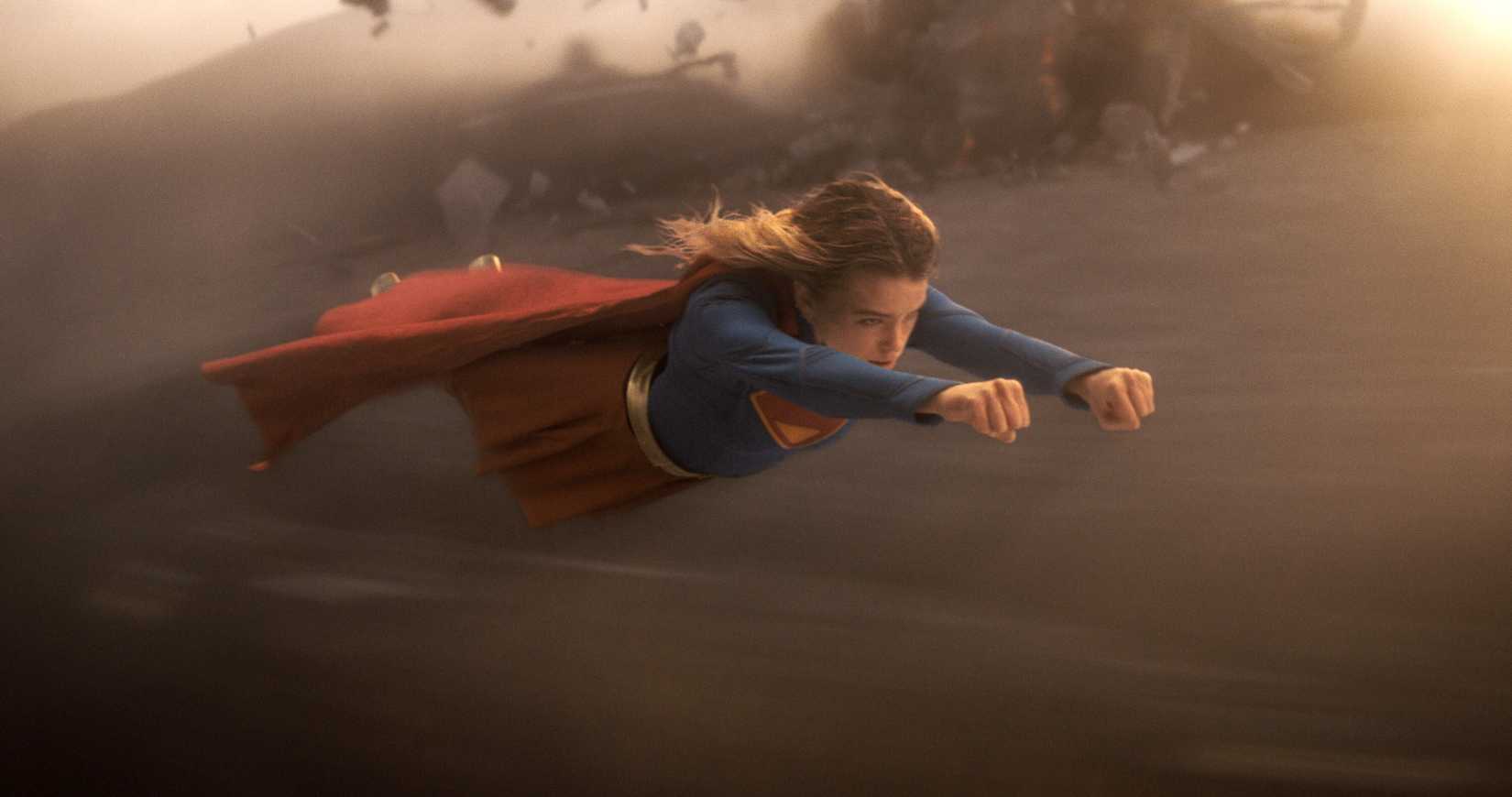 Milly Alcock as Supergirl, flying through a sandy landscape, fists clenched ahead of her