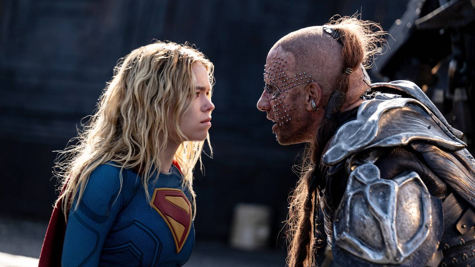 Milly Alcock as Supergirl, in the traditional costume, faces off against Krem, a man wearing patched-together metal armor, a shaved head and long topknot ponytail, and an extensive grid of facial piercings