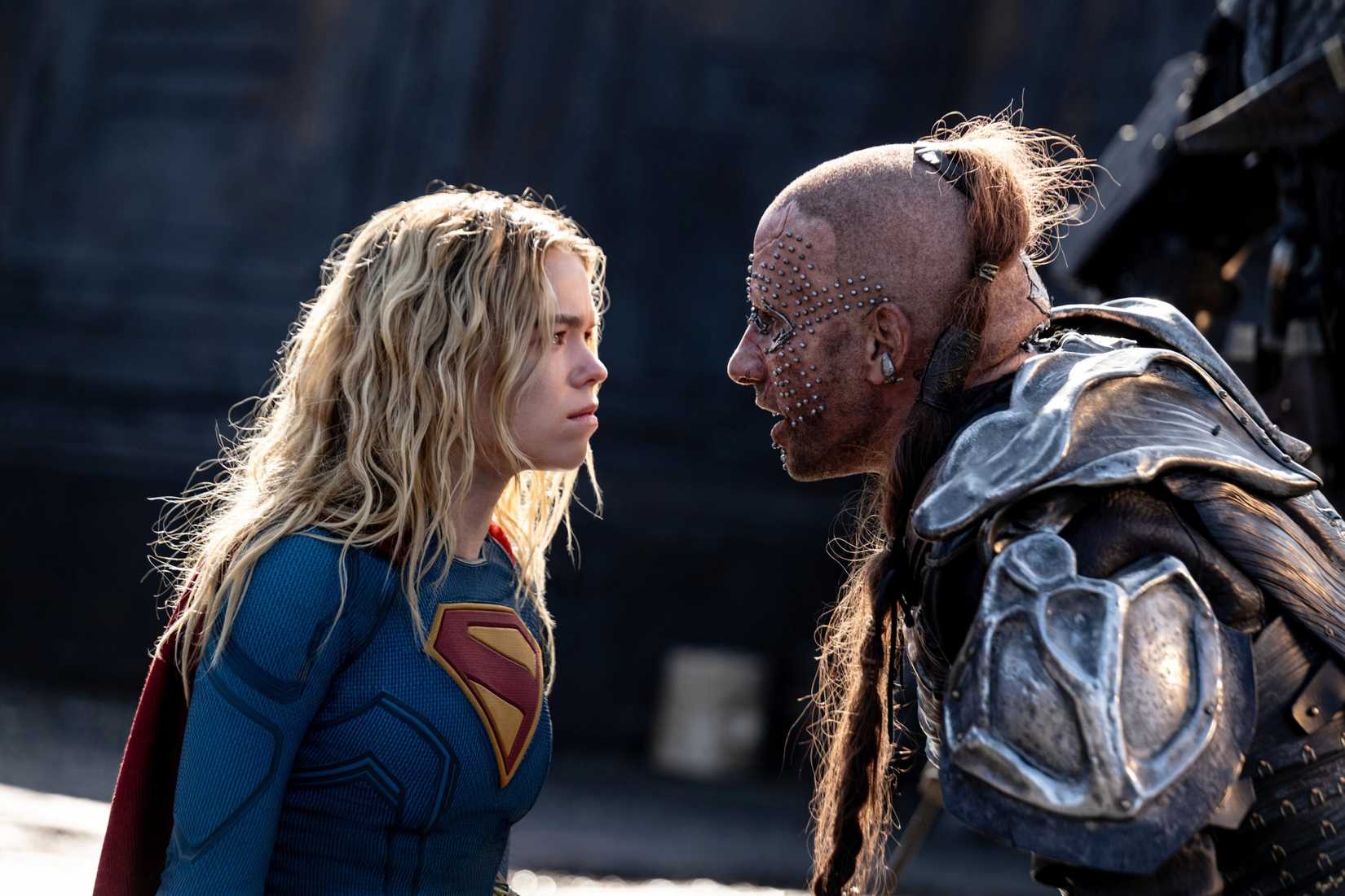 Milly Alcock as Supergirl, in the traditional costume, faces off against Krem, a man wearing patched-together metal armo