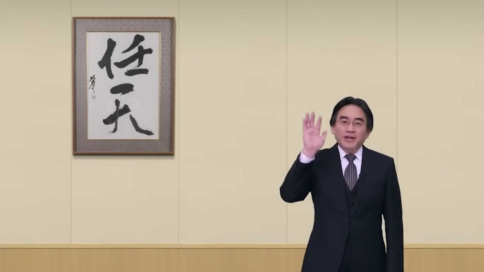 Satoru Iwata waves in front of a blank wall with framed Japanese calligraphy hanging on it
