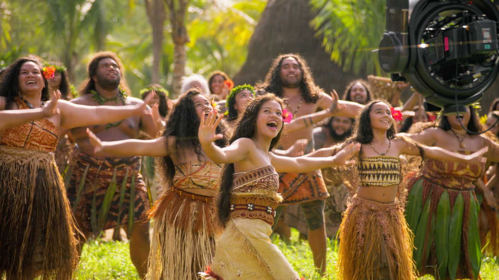 Moana (Catherine Laga'aia) and a crowd of actors portraying Pacific Island villagers dance together in the 2026 live-action Moana, with a wire-covered camera in frame