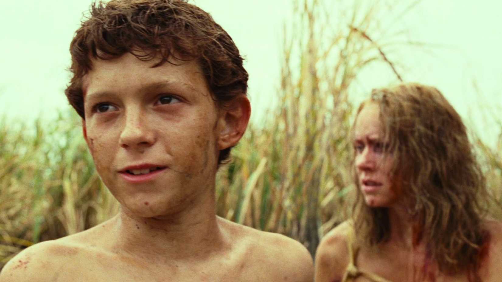 Tom Holland, dirtied and grim looking, looks ahead while Naomi Watts stares off in the background.