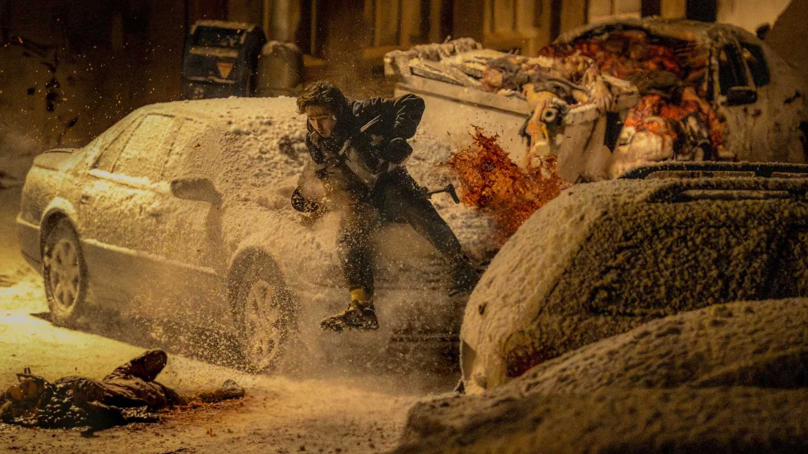 Bryan (Austin Abrams) runs past snow covered cards as bodies fall near him in Resident Evil