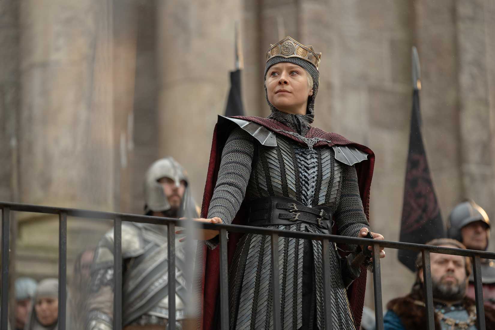 Queen Rhaenyra Targaryen (Emma D'Arcy) looks over a railing while her guards and council stand behind her in House of the Dragon