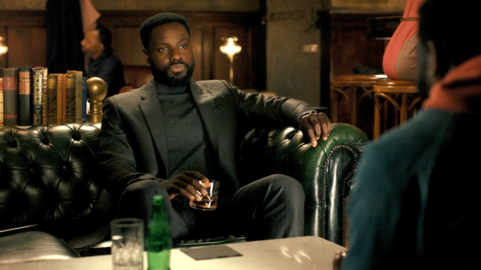 Leighton Vance (Dayo Okeniyi) sits on a leather couch and holds a drink while talking to another version of himself in Dark Matter season 2