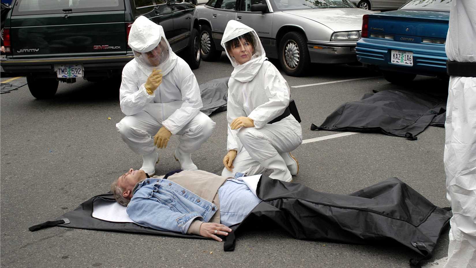 Two figures wearing hazmat suits squat near a man in a body bag in The 4400
