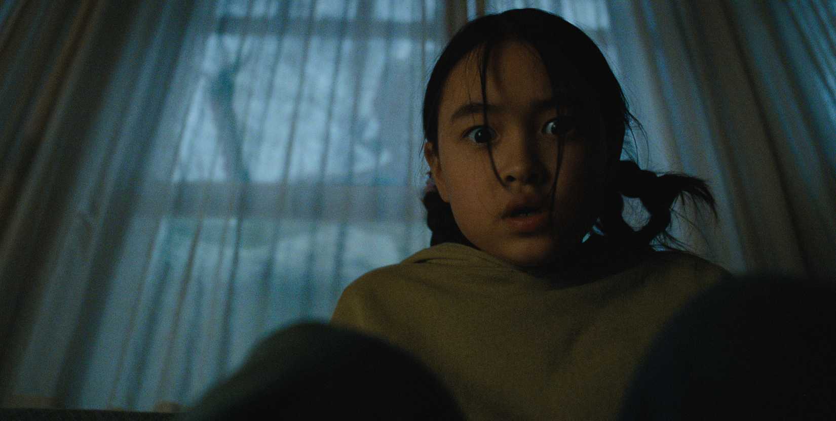 Ruth, a young girl, stares directly into the camera as a shadowy figure leans against the window.