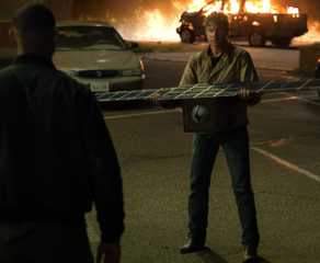Hal Jordan (Kyle Chandler) holds out a satelite to John Stewart (Aaron Pierre) as a pickup truck burns in the background in Lanterns