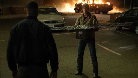 Hal Jordan (Kyle Chandler) holds out a satelite to John Stewart (Aaron Pierre) as a pickup truck burns in the background in Lanterns