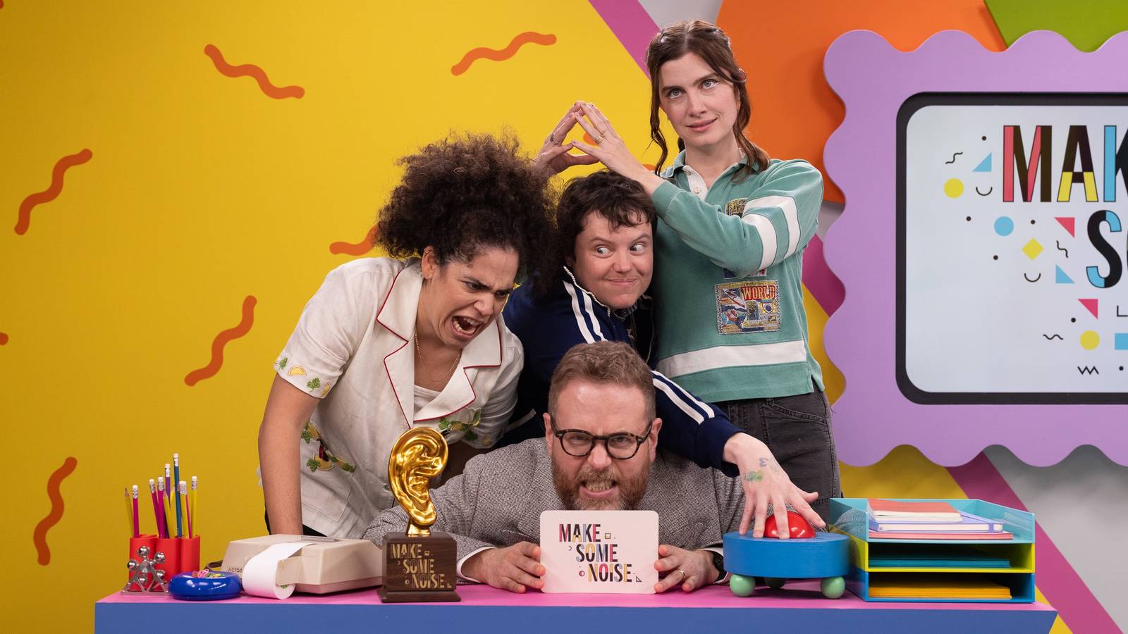 Make Some Noise photo showing three contestants (Vic Michaelis, Ally Beardsley, and Kimia Behpoornia) standing over host Sam Reich, who's sitting at a desk.