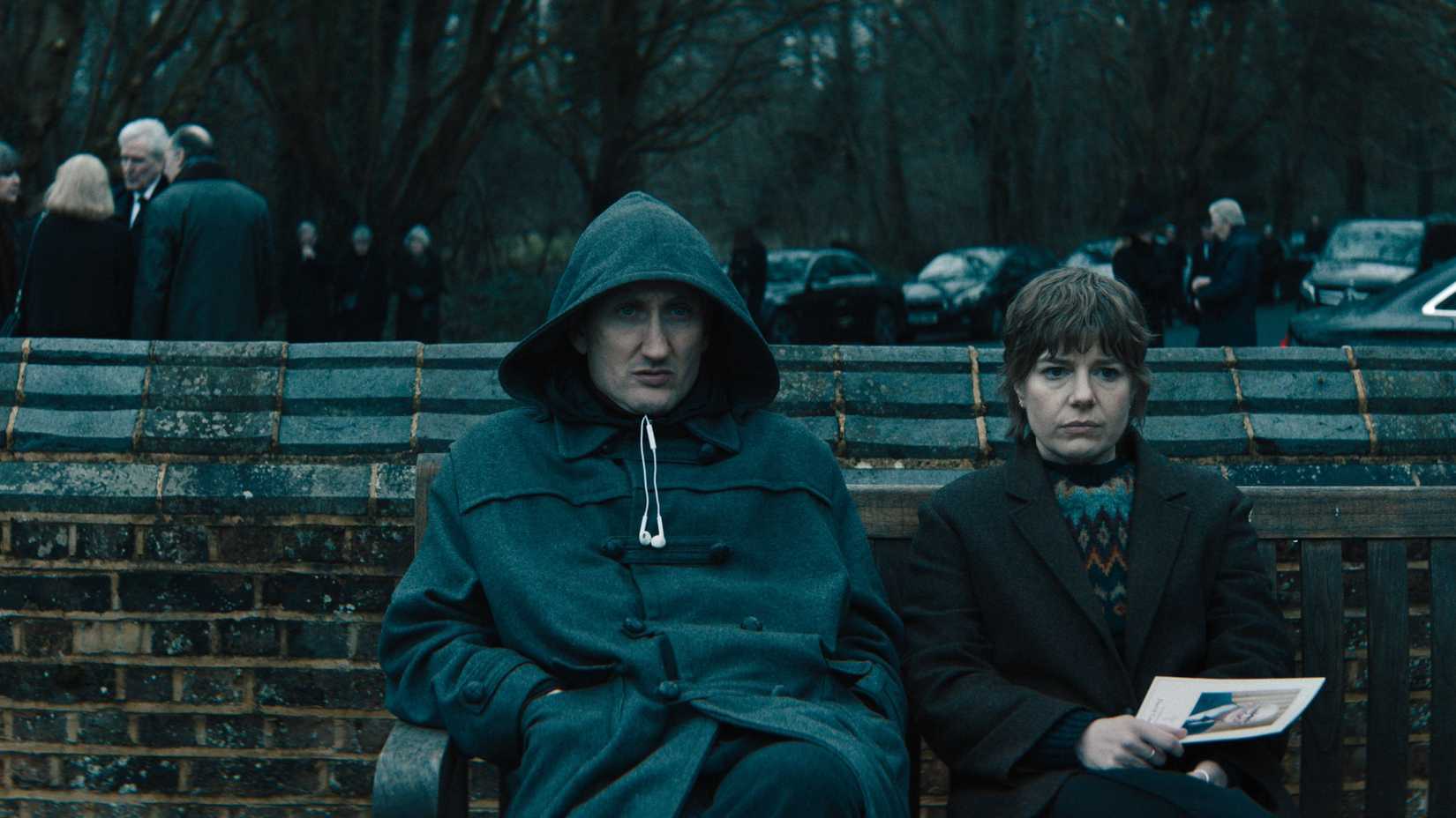 J.K. Coe (Tom Brooke) sits on a bench next to Shirley Dander (Aimee-Ffion Edwards) who holds a funeral program in Slow Horses season 6.