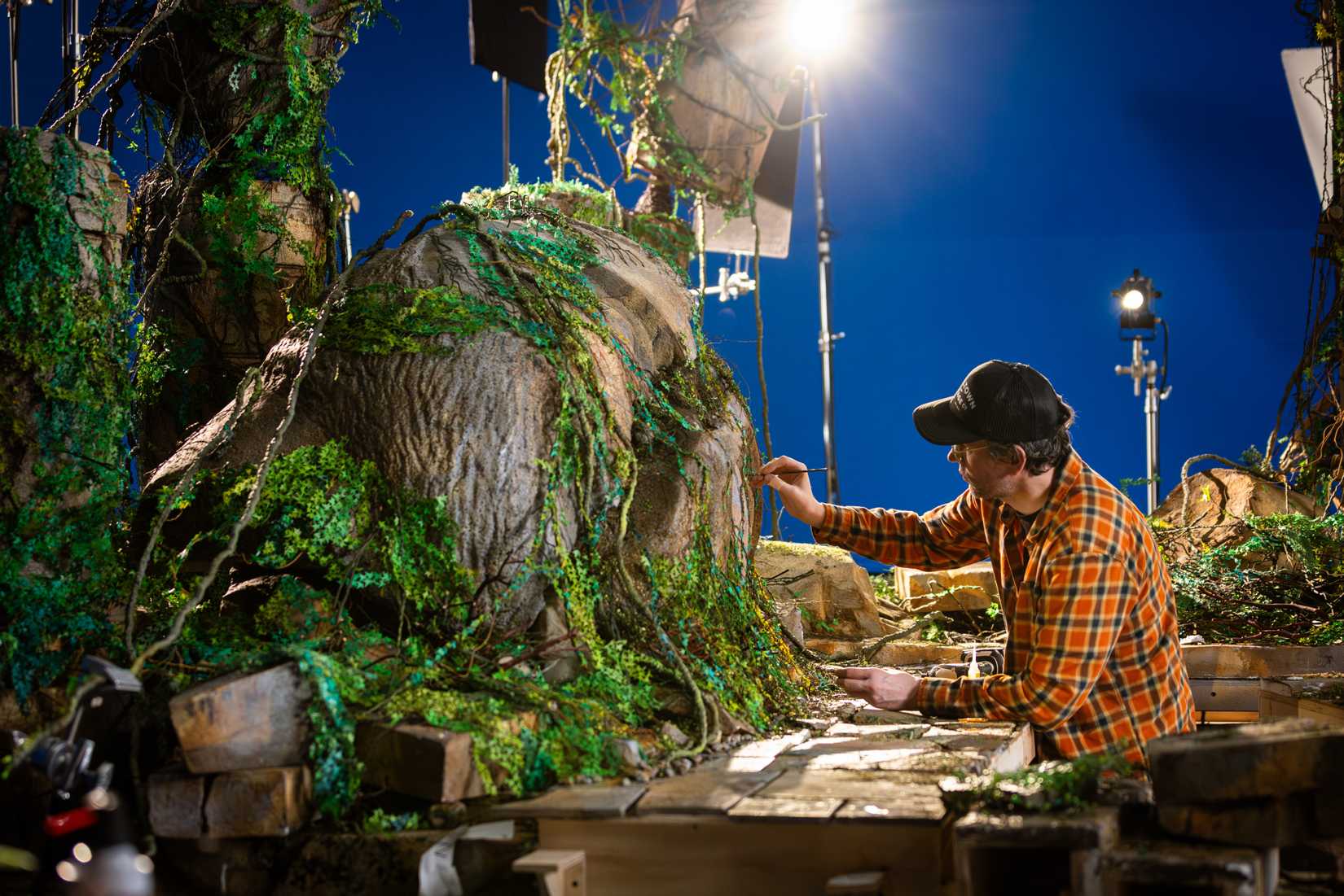 Behind the scenes of Wildwood, a man in flannel painting the side of a cliff with a blue screen in the background