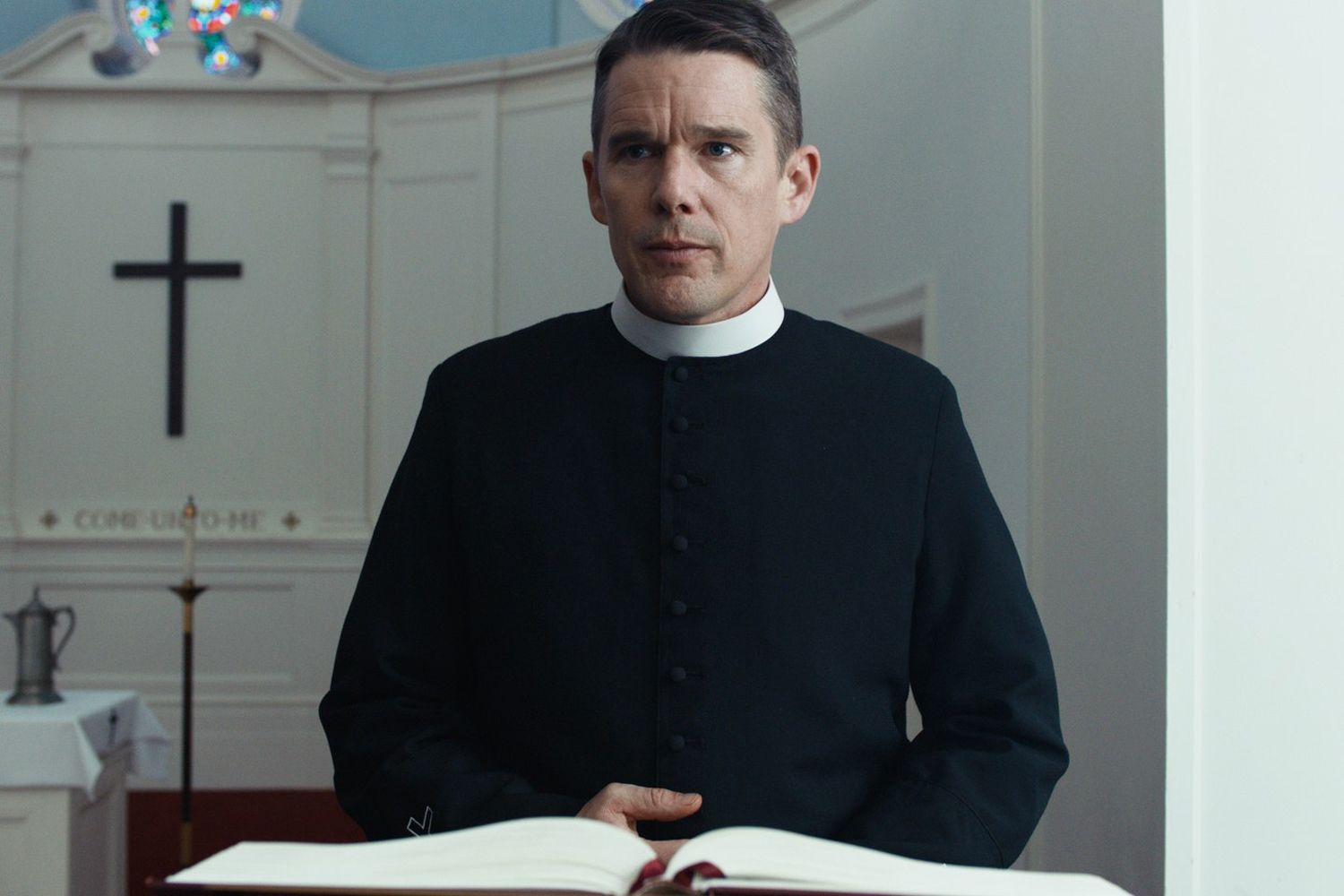 A man in priest attire (Ethan Hawke) stands behind a lectern with a black cross visible in the background.