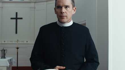 A man in priest attire (Ethan Hawke) stands behind a lectern with a black cross visible in the background.