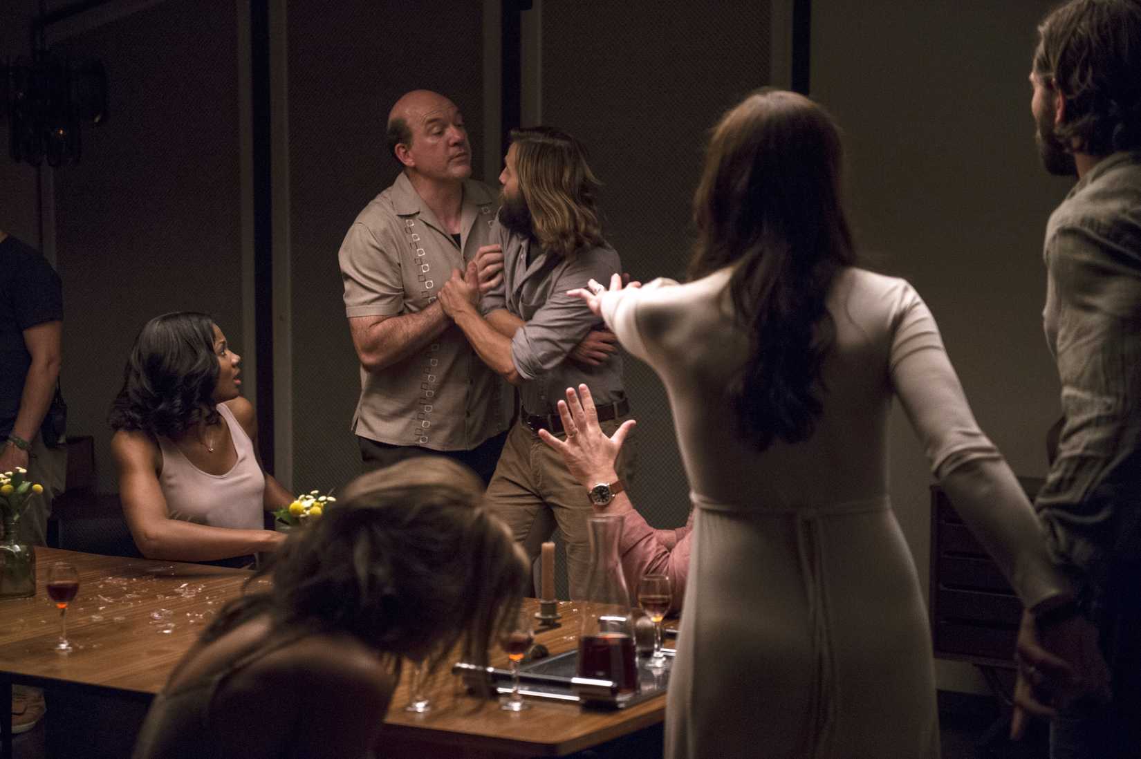 In a scene from the 2016 film The Invitation, a group of people sits and stands around a dinner table looking contentious and confused; an older man grabs ahold of a younger woman while another woman with her back to the camera holds out her hand as if trying to stop them.
