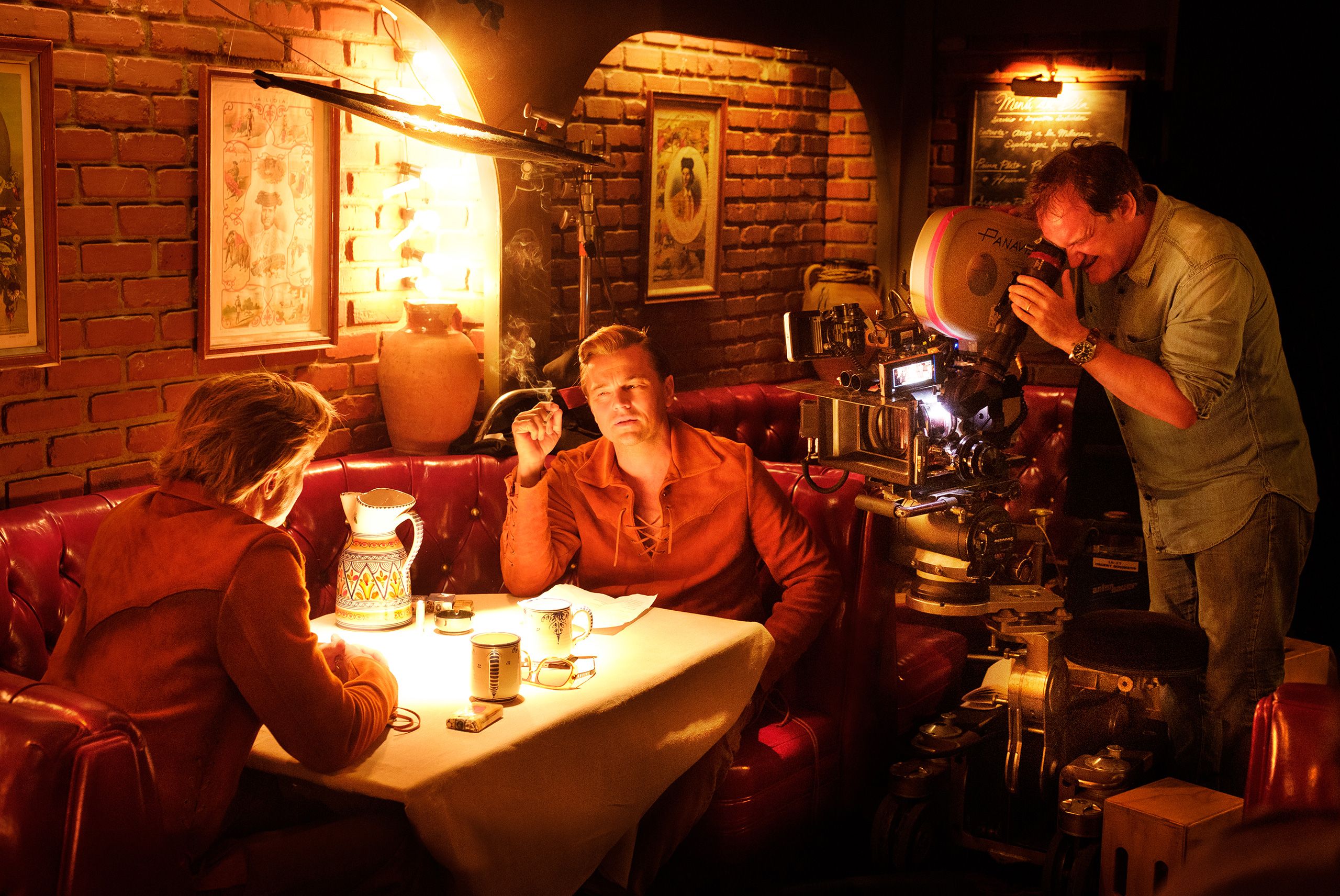 Brad Pitt and Leonardo DiCaprio sit facing each other in a restaurant booth as Quentin Tarantino watches from behind the camera filming Once Upon a Time ... in Hollywood
