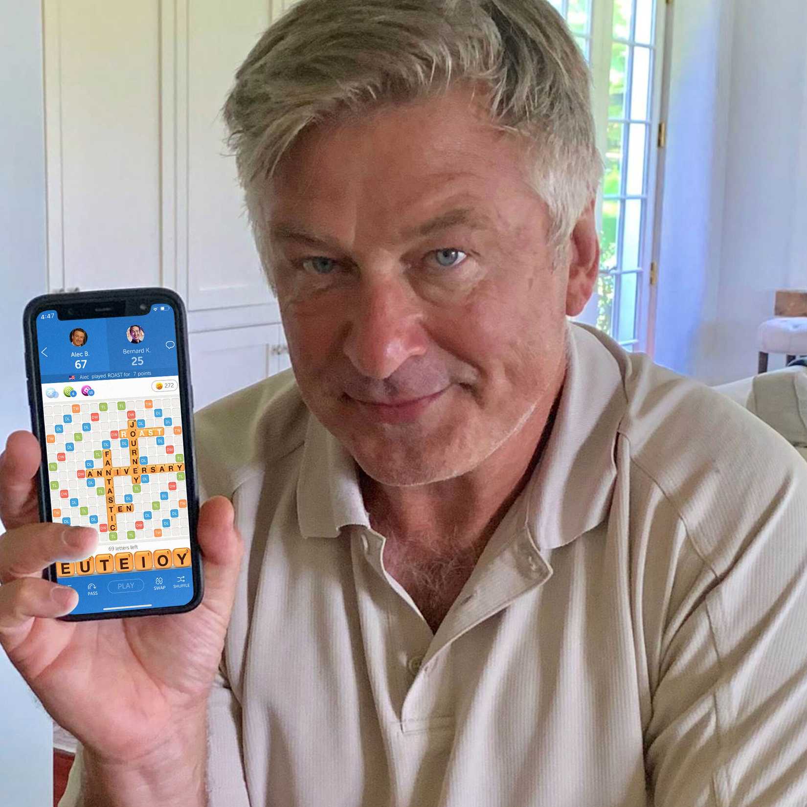 An aging man — Alec Baldwin — with gray hair and a tan polo shirt has a mostly neutral expression on his face. He is slightly smirking and holding up an iPhone with a game of Words With Friends on the screen.