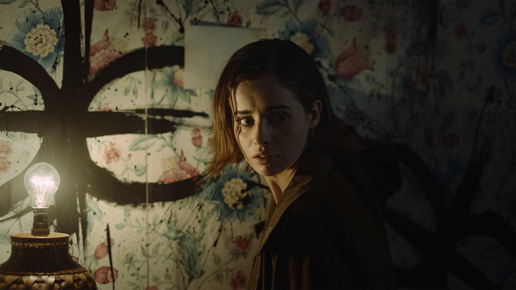 Erica, a white woman with dark hair, stands in a dimly lit room. She is looking over her shoulder with a concerned expression. A symbol has been painted on the wall.
