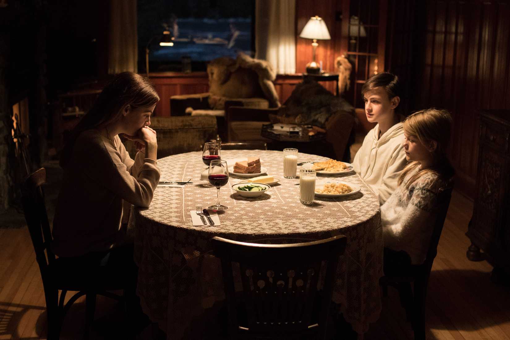 A woman sits across from two children at a brightly lit dining-room table in a very dark room in The Lodge