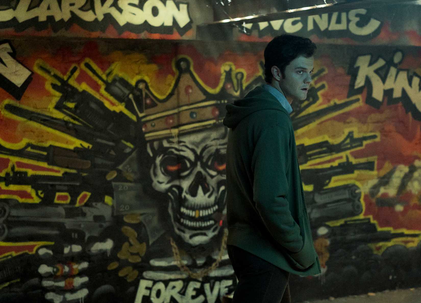 Jack Quaid as Hughie in The Boys, passing in front of a mural of a crowned skull wreathed in guns