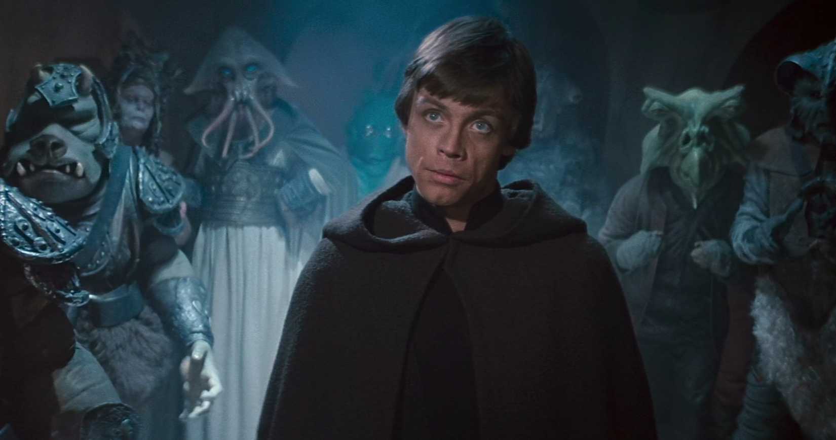 Luke Skywalker stands in Jabba’s Palace, wearing a hooded black cloak, with a crowd of non-humans behind him in Star Wars: Return of the Jedi
