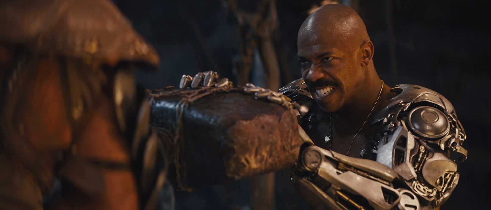 Jax (Mehcad Brooks), a Black man with a shaved head and powerful mechanical arms, grimaces as he grips a large brown block in 2021's Mortal Kombat