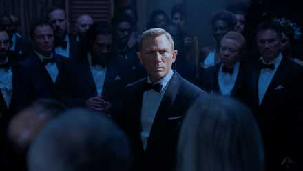 Daniel Craig as James Bond in No Time to Die, standing in a spotlight, surrounded by formal-suited members of SPECTRE