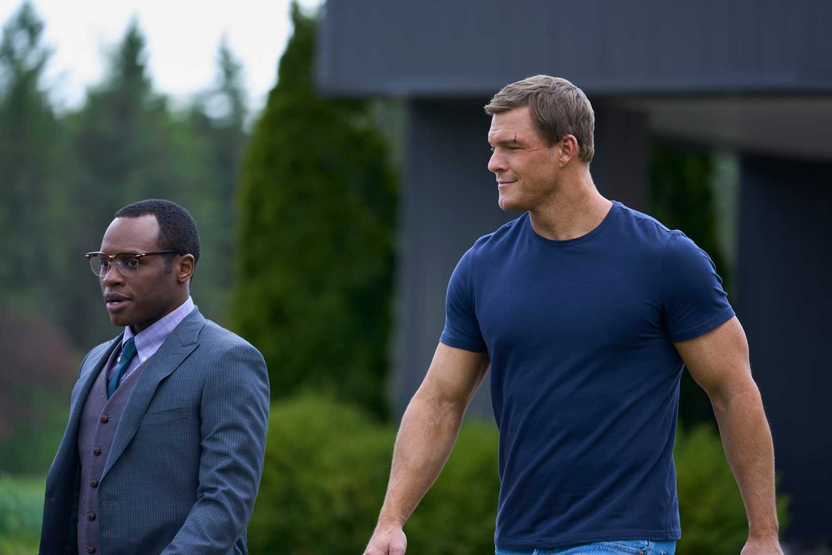 Reacher, a very large man in a small-fitting T-shirt, walks next to a much smaller man in a three-piece suit