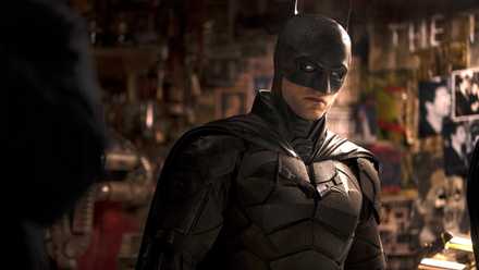 Robert Pattinson as Batman standing in front of a Riddler riddle wall